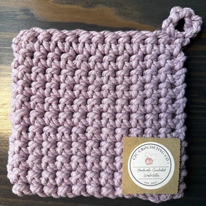 Op de afbeelding: Een paarse gehaakte ovenwant met een lusvormige handgreep en een bruin label met de tekst "GIC CROCHETING CO. Handmade Crocheted Washcloths - EST. 2023"