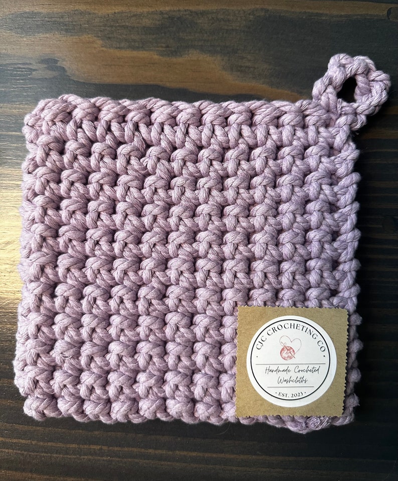 Pu&ograve; includere: Un sottopentola di colore viola lavorato a uncinetto con un manico ad anello e un'etichetta marrone che dice "GIC CROCHETING CO. Handmade Crocheted Washcloths - EST. 2023"