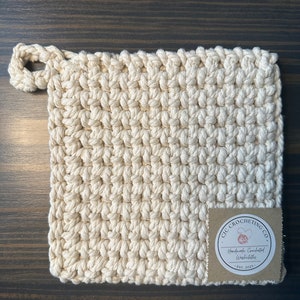 May include: A cream-colored crocheted pot holder with a loop handle. The pot holder has a brown label with the text "CIC CROCHETING CO. Handmade Crocheted Washcloths Est. 2021."