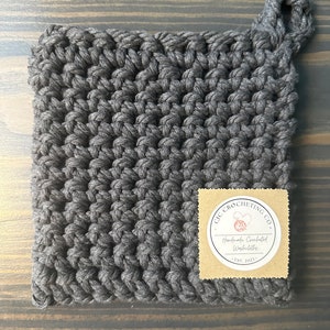 Peut inclure: Un dessous de plat en crochet gris avec une anse en boucle. Le dessous de plat porte une étiquette en papier brun avec le texte "QIC CROCHETING CO. Handmade Crocheted Washcloths Est. 2021."
