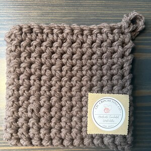 Könnte beinhalten: Ein brauner gehäkelter Topflappen mit einer Schlaufe. Ein kleines braunes Papieretikett mit einem weißen Kreis und dem Text "CIC CROCHETING CO. Handmade Crocheted Washcloths EST. 2021" ist am Topflappen befestigt.