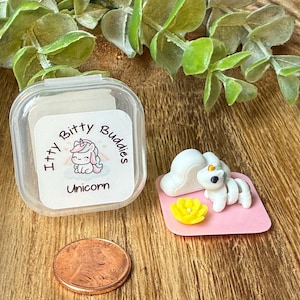 May include: A small clear plastic container with a label reading Itty Bitty Buddies Unicorn, a miniature white unicorn figurine with a yellow flower on a pink square base, and a copper penny for scale, all displayed on a wooden surface.