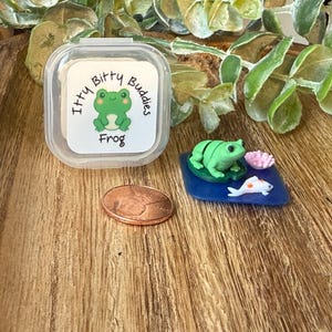 May include: A clear plastic container with a sticker that reads "Itty Bitty Buddies Frog" next to a miniature frog figurine on a blue base with a koi fish and a pink flower. A penny is also in the image.