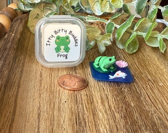 May include: A clear plastic container with a label reading Itty Bitty Buddies Frog, a copper penny, and a miniature green frog figurine on a blue lily pad with a small white fish and a pink flower. The items are arranged on a wooden surface.