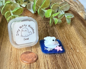 May include: A small white seal figurine resting on a blue square base with a tiny silver fish and a pink starfish. Next to it is a clear plastic container labeled Itty Bitty Buddies Seal and a copper penny for scale on a wooden surface.
