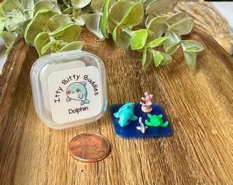 May include: A clear plastic container with a label reading Itty Bitty Buddies Dolphin, next to a copper penny and a blue resin square featuring a miniature blue dolphin, a green sea turtle, a small grey shark, and a pink coral piece.