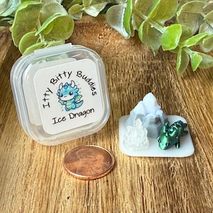 May include: A clear plastic container with a label reading Itty Bitty Buddies Ice Dragon, a small green dragon figurine, a translucent crystal-like piece, a grey mountain-shaped object on a white base, and a copper penny for scale.