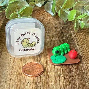 May include: A miniature green caterpillar figurine with a small red apple and a green leaf on a brown square base, next to a copper penny and a clear plastic container with a label reading Itty Bitty Buddies Caterpillar in black text.