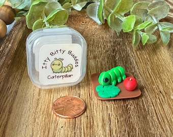 Könnte beinhalten: Eine Miniatur-Raupenfigur in Grün mit einem kleinen roten Apfel und einem grünen Blatt auf einem braunen quadratischen Sockel, neben einem durchsichtigen Kunststoffbehälter mit der Aufschrift Itty Bitty Buddies Caterpillar und einer Kupfermünze.