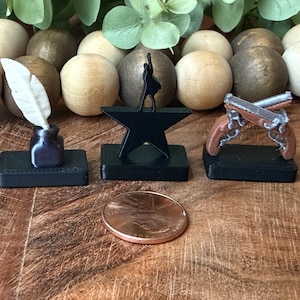 May include: Three miniature figurines on black rectangular bases: a white quill in a dark inkwell, a black star with a silhouette of a person standing on top, and two crossed brown and silver pistols. A copper penny is placed in front for scale.