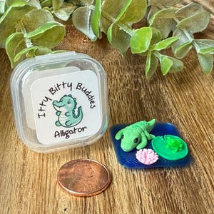 May include: A clear plastic container with a label reading Itty Bitty Buddies Alligator, a copper penny for scale, and a miniature green alligator figurine on a blue square base with a small green lily pad and a pink flower.