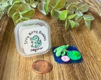 May include: A clear plastic container with a label reading Itty Bitty Buddies Alligator, a copper penny for scale, and a miniature green alligator figurine on a blue square base with a small green lily pad and a pink flower.