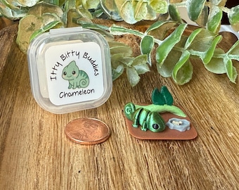 May include: A small green articulated chameleon figurine on a brown base with a tiny gray food bowl and a green leaf, next to a copper penny and a clear plastic container with a label reading Itty Bitty Buddies Chameleon in black text.