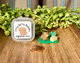 May include: A clear plastic container with a label reading Itty Bitty Buddies Highland Cow, a miniature brown Highland cow figurine on a green base with a small hay bale and green grass, and a copper penny for scale on a wooden surface.