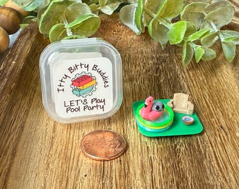 May include: A clear plastic container with a label reading Itty Bitty Buddies LET'S Play Pool Party, a copper penny, and a miniature green base featuring a small inflatable pool with a pink flamingo float, a tiny gray figure, and a lounge chair.