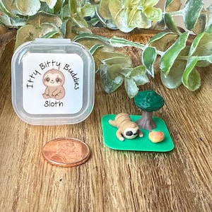 May include: A clear plastic container with a label that reads "Itty Bitty Buddies Sloth" next to a miniature sloth figurine, a tree, and a small orange fruit on a green base. A penny is also in the image.