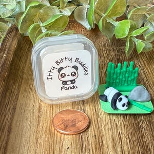 May include: A clear plastic square container with a white label reading Itty Bitty Buddies Panda, a miniature black and white panda figurine on a green base with a green bamboo stalk and a gray rock, and a copper penny for scale.