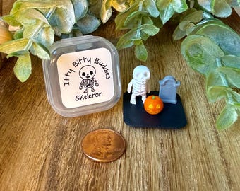 May include: A clear plastic container with a label reading Itty Bitty Buddies Skeleton, accompanied by a miniature white skeleton figurine, a small orange jack-o'-lantern, and a gray tombstone on a black base, next to a copper-colored penny.