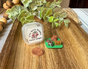 May include: A clear plastic container with a label reading Itty Bitty Buddies Forest Dragon, a small green square base with a miniature metallic dragon, a brown tree stump, and a gold treasure chest, and a copper penny for scale.
