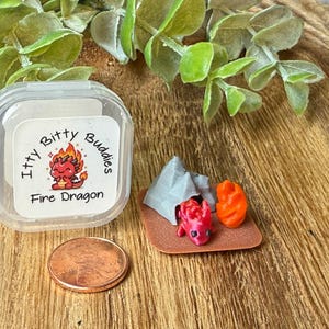 May include: A miniature set featuring a small red dragon, a gray tent, and an orange campfire on a brown base, next to a clear plastic container labeled Itty Bitty Buddies Fire Dragon and a copper penny for scale on a wooden surface.