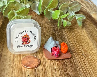 May include: A miniature set featuring a small red dragon figurine, a grey tent, and an orange campfire on a brown base, next to a clear plastic container labeled Itty Bitty Buddies Fire Dragon and a copper penny for scale reference.