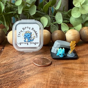 May include: A miniature blue dragon figurine with a yellow lightning bolt accessory and a gray cave structure, displayed next to a clear plastic container labeled Itty Bitty Buddies Lightning Dragon and a copper penny for scale.