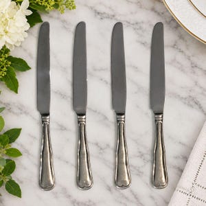 May include: A set of four silver-toned stainless steel dinner knives arranged vertically. Each knife features a smooth, rounded handle with a subtle heart-shaped detail at the base and a polished, reflective blade with a slightly serrated edge.