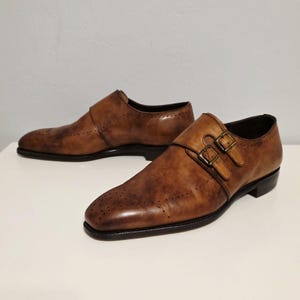 May include: A pair of brown leather shoes with a double monk strap fastening. The shoes feature a classic design with decorative perforations on the toe and sides. The leather has a distressed finish, giving them a vintage appearance.