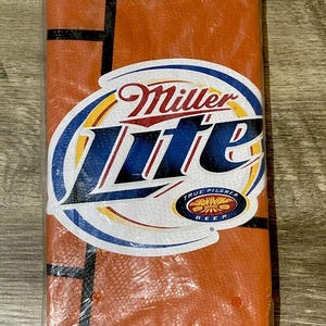 Könnte beinhalten: Orange-schwarzes Handtuch im Basketball-Design in einer transparenten Plastiktüte. Das Handtuch hat schwarze Linien, die einem Basketballfeld ähneln. Das Miller Lite-Logo ist mit den Worten "True Pilsner Beer" abgebildet.