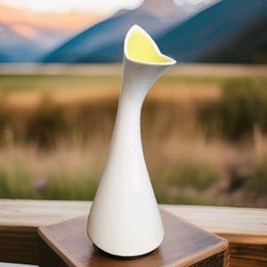 以下が含まれることがあります： 白のセラミック花瓶。開口部はフレアで非対称、内側は黄色です。花瓶は細長く、細長い形状で、木製の表面に立っています。背景には、山と緑のあるぼやけた風景が写っています。