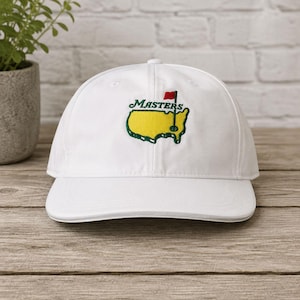May include: A white baseball cap featuring an embroidered logo on the front. The logo displays a yellow map of the United States with a green outline, the word MASTERS in green text, and a small red flag on a green pole positioned over the map.
