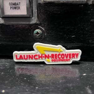 May include: White plastic patch with a red and yellow logo. The logo is a stylized yellow lightning bolt with the text "LAUNCH-N-RECOVERY EQUIPMENT" in red letters.