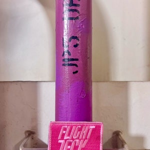 May include: A pink rectangular patch with the text "FLIGHT TECK" in white lettering. The patch is placed below a purple painted pipe with black text. The background is a light beige color.