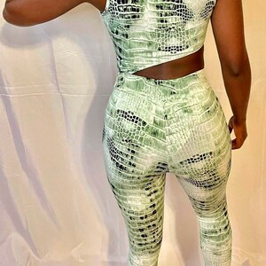 以下が含まれることがあります： 背中がカットアウトされた、緑と黒のヘビ柄のジャンプスーツ。 ノースリーブのデザインで、体にフィットしたシルエットです。