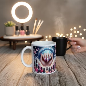 Peut inclure: Mug en céramique blanche avec un motif de nail art. Le motif représente une main tenant un globe lumineux avec de longs ongles colorés, entourée de flacons de vernis à ongles. Un mug noir et une lampe annulaire sont en arrière-plan.