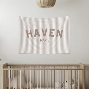 May include: A striped wall tapestry with the name "HAVEN" and the name "MARIE" in a soft brown color. The tapestry hangs above a wooden crib in a nursery setting. The room has a neutral color palette.
