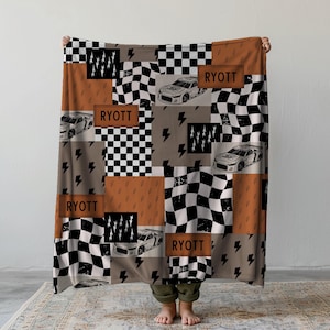 May include: A patchwork blanket featuring race cars, lightning bolts, and checkered flags in shades of brown, gray, and black. The name "RYOTT" is displayed on several patches. The blanket is held up by a person.