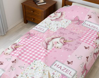 May include: Pink patchwork quilt with unicorns, butterflies, and bows. The quilt has the name "HAZEL" on a label. The design includes floral and gingham patterns. The quilt is on a bed.
