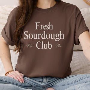 Könnte beinhalten: Ein braunes T-Shirt mit den Worten "Fresh Sourdough Club" in einer Serifenschrift. Unter dem Text stehen die Wörter "Fold" und "Rise". Das Shirt wird von einer Person getragen, die auf einem Bett sitzt.
