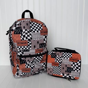 May include: A backpack and matching lunch bag set. The set features a patchwork design with black and white checkered squares, orange rectangles, gray sections, and lightning bolt graphics. The name "ROMAN" is printed on several patches.