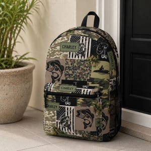 May include: A backpack featuring a camouflage and fishing-themed pattern with black, green, and tan colors. The design includes illustrations of fish, fishing hooks, and a person in a boat, with the name CHARLEY printed on several green labels.