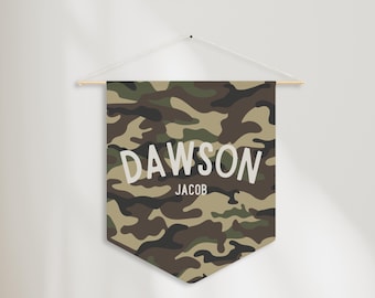 Può includere: Un banner da parete con motivo mimetico con il nome "DAWSON" e il nome "JACOB" in lettere bianche. Lo striscione è appeso a un'asta di legno e a una corda.