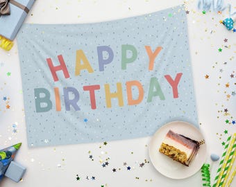 May include: A light blue tapestry with the words "HAPPY BIRTHDAY" in colorful letters. The tapestry is decorated with small white and gold speckles. A slice of cake sits on a white plate, surrounded by party decorations.