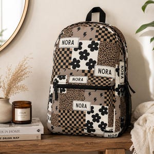 May include: A backpack featuring a patchwork design of black and white flowers, brown and black checkerboard patterns, leopard print, and lightning bolts. The name NORA is printed on multiple white rectangular patches across the front and sides.
