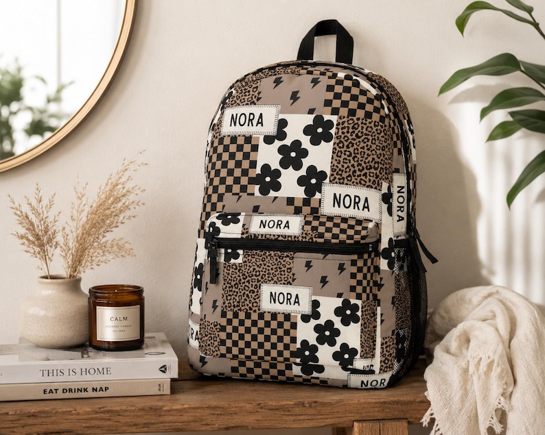 May include: A backpack featuring a patchwork design of black and white flowers, brown and black checkerboard patterns, leopard print, and lightning bolts. The name NORA is printed on multiple white rectangular patches across the front and sides.