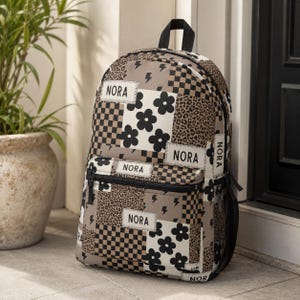 May include: A backpack featuring a patchwork design of beige, black, and white patterns including leopard print, checkerboard, lightning bolts, and flowers. The bag has multiple rectangular patches with the name NORA printed in black text.