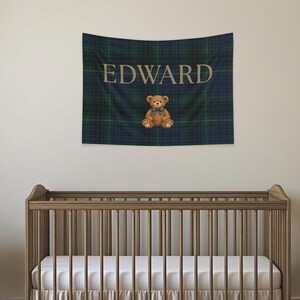 May include: A tapestry with the name "EDWARD" in tan lettering over a blue and green plaid pattern. A brown teddy bear with a blue bow tie sits below the name. A wooden crib is in the foreground.