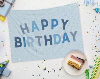 May include: A light blue banner with the words "HAPPY BIRTHDAY" in shades of blue. The banner is decorated with small white and blue specks. A slice of cake sits on a white plate, surrounded by party decorations.