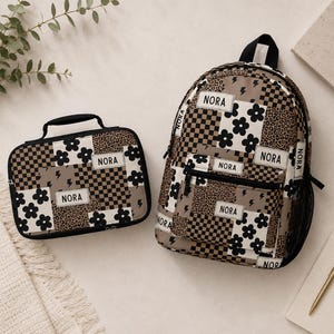 May include: A backpack and a rectangular lunch bag featuring a patchwork design of black and white flowers, brown and black checkerboard patterns, leopard print, and lightning bolts. Both items have white rectangular labels with the name NORA.