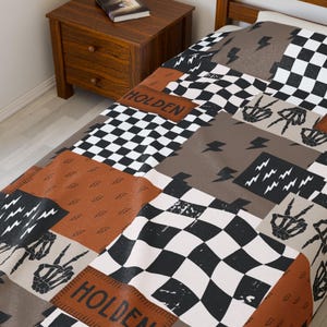May include: A patchwork-style blanket with a bold design. The blanket features squares of black and white checkerboard, brown, and gray with lightning bolts and skeleton hand graphics. The word "HOLDEN" is printed on a brown rectangle.